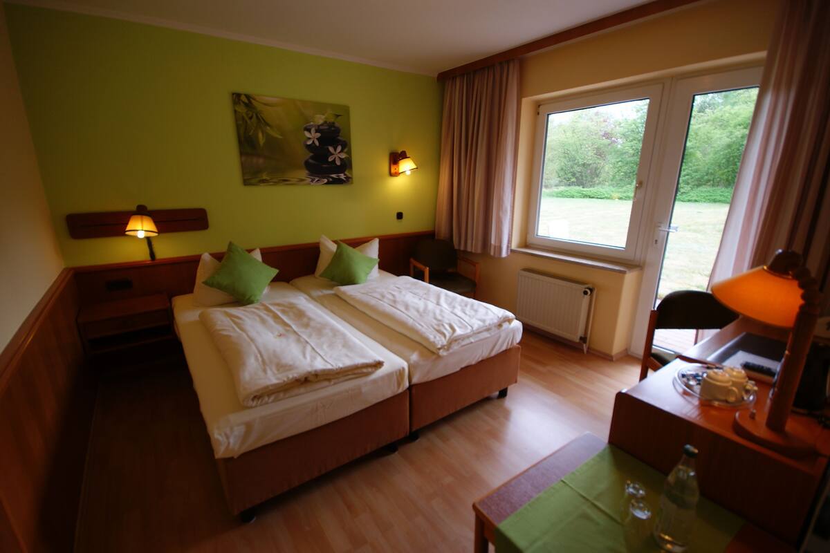A hotel room with a double bed, two bedside lamps, a desk with a lamp, a chair, a window with curtains, and a painting on the wall.