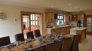 Un coin repas avec une longue table dressée pour le repas, des meubles de rangement en bois et une cuisine en arrière-plan.