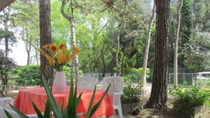 An outdoor dining area with a table set for two, surrounded by trees and greenery. - Charming apartment with fenced garden (Bibione)