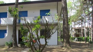 A building with a white exterior and blue shutters, surrounded by trees and a paved walkway. - Charming apartment with fenced garden (Bibione)