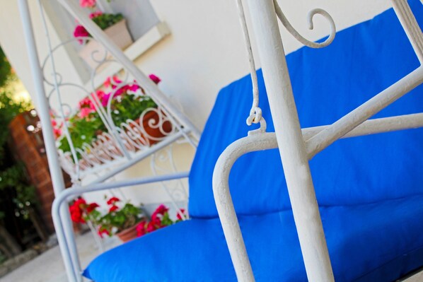 A blue chair with a metal frame, positioned outdoors with a blue wall and potted plants in the background.