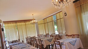 Un espace repas comprenant plusieurs tables et chaises, de grandes fenêtres avec des rideaux et un lustre.