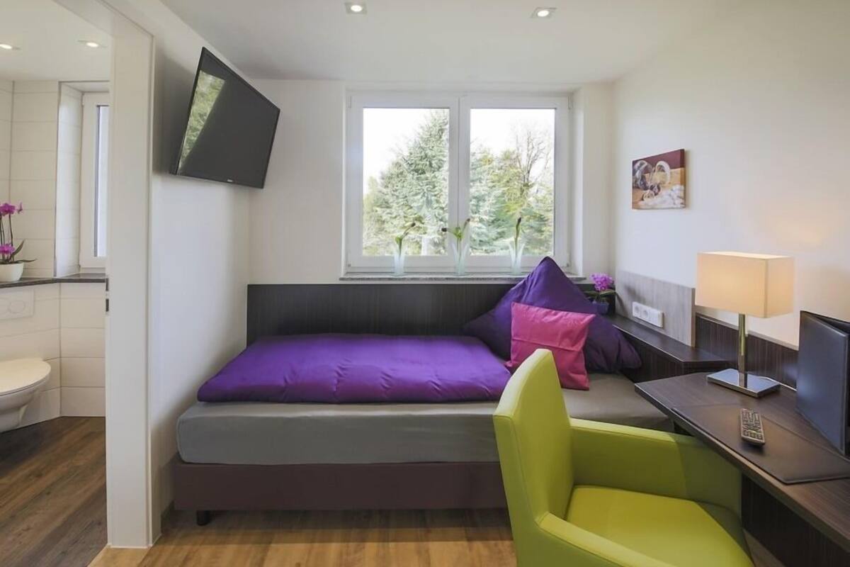 A modern room with a bed, a desk, a chair, a TV, and a window with a view of trees.
