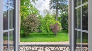 Vista a través de una ventana con barandilla de hierro forjado, que da a un jardín cuidado con varios árboles y césped.