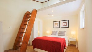Un dormitorio con una escalera de madera, una cama con una manta roja, dos lámparas de noche y cuadros enmarcados en la pared.