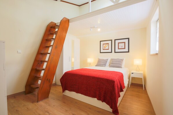 A bedroom with a wooden staircase, a bed with a red blanket, two bedside lamps, and framed pictures on the wall.