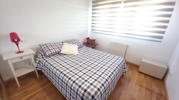 A bedroom with a bed, a nightstand, a lamp, a window with blinds, and a radiator.