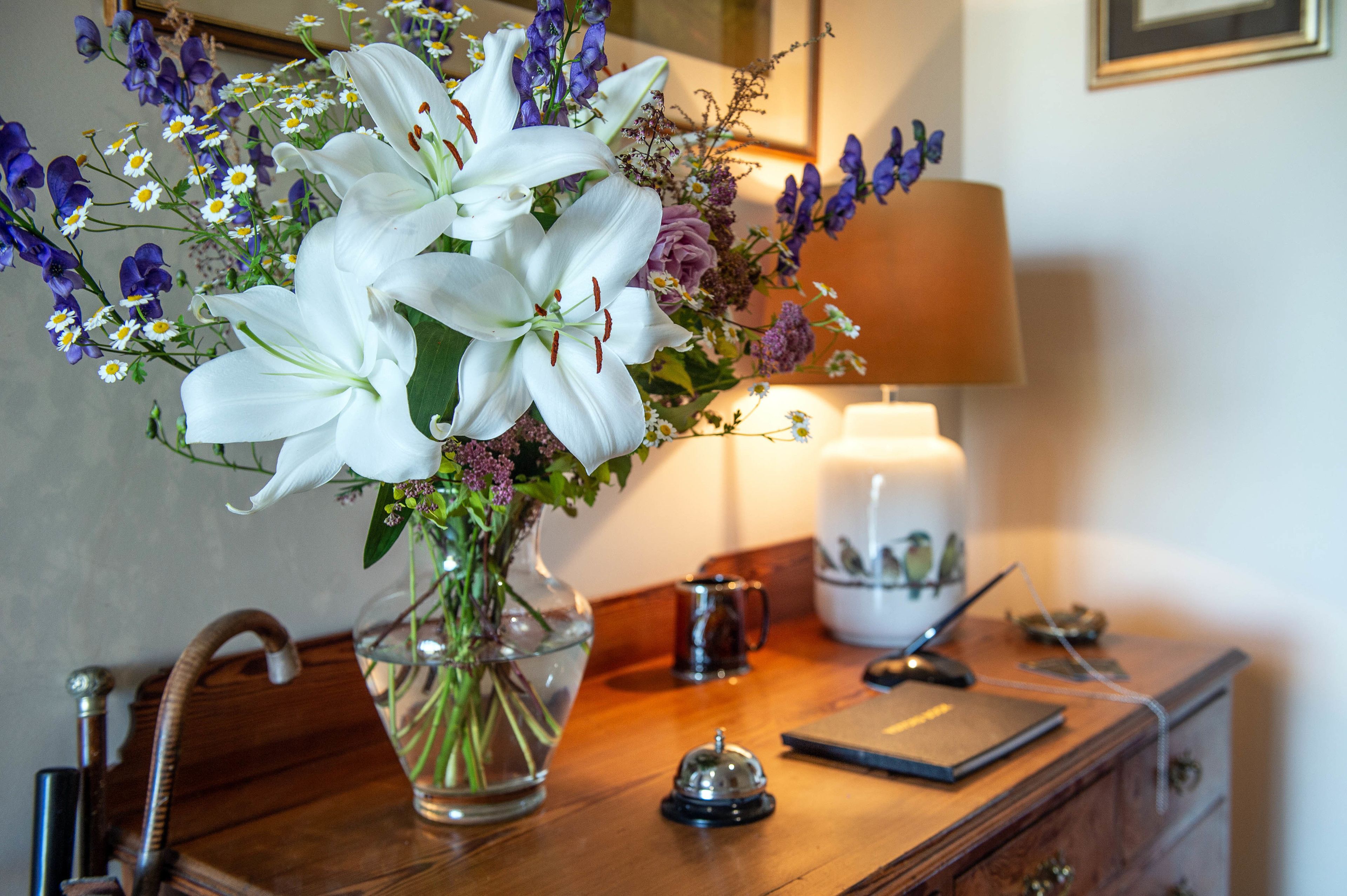 Een houten bureau met een transparante glazen vaas gevuld met witte lelies en paarse bloemen, een lamp, een koffiemok en een kleine bel.
