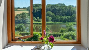 Janela com vista para um lago e vegetação, um vaso com flores, e uma pequena mesa com bule e xícara.