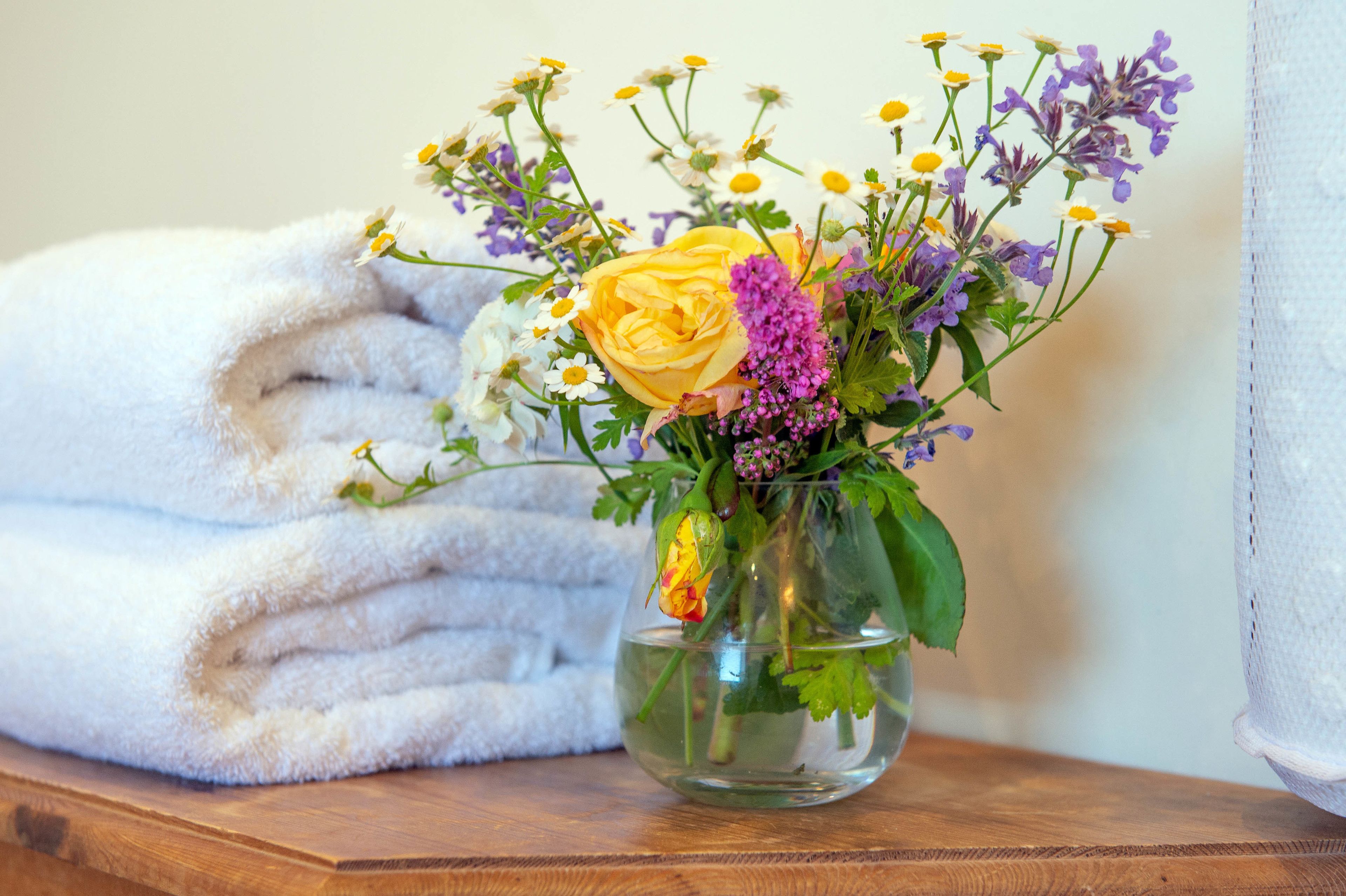 A clear glass vase with a bouquet of flowers and a folded white towel on a wooden surface.