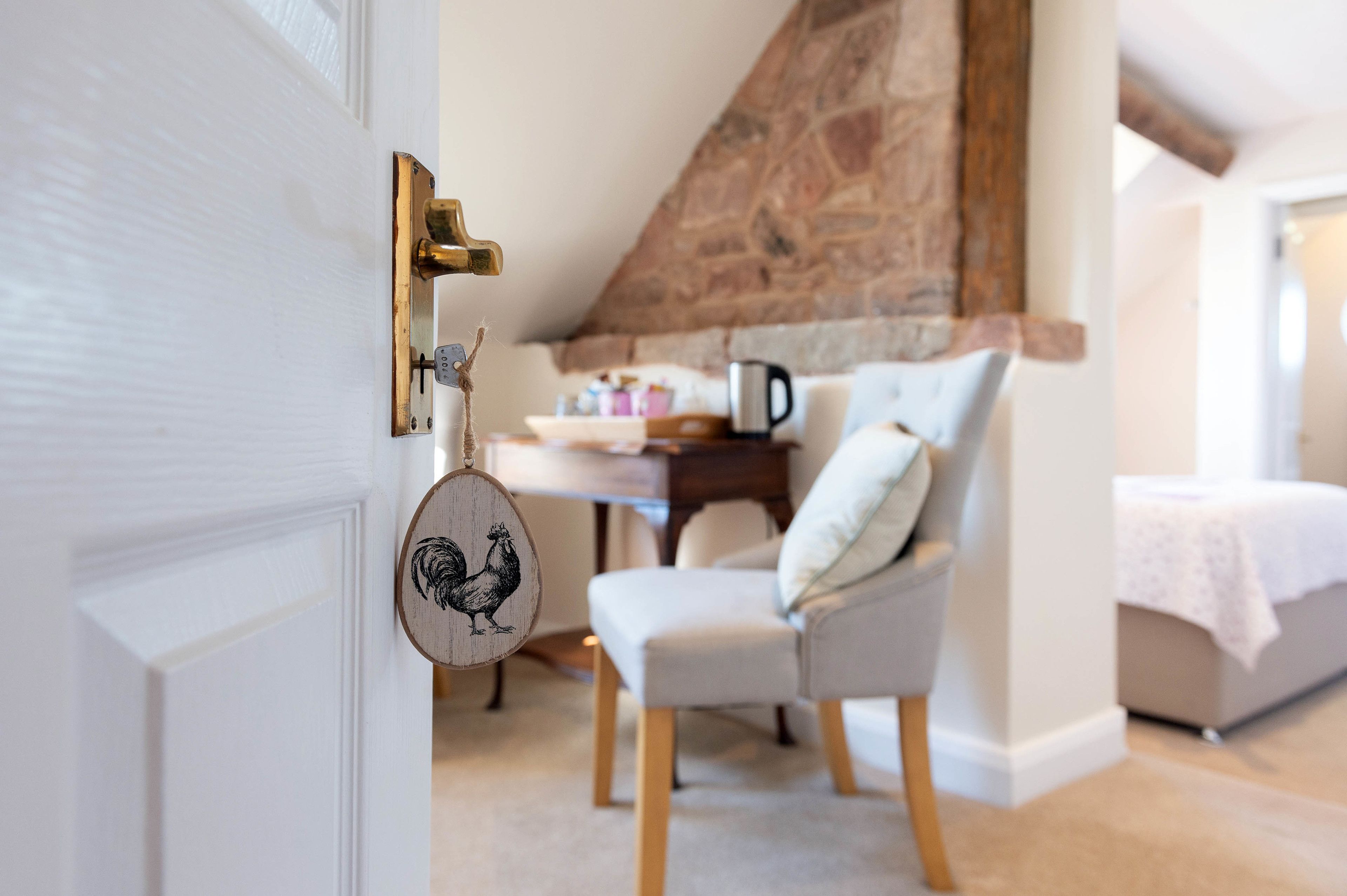 A cozy room with a stone fireplace, a white door with a brass handle, a wooden rooster decoration, a grey armchair, and a bed with a white cover.