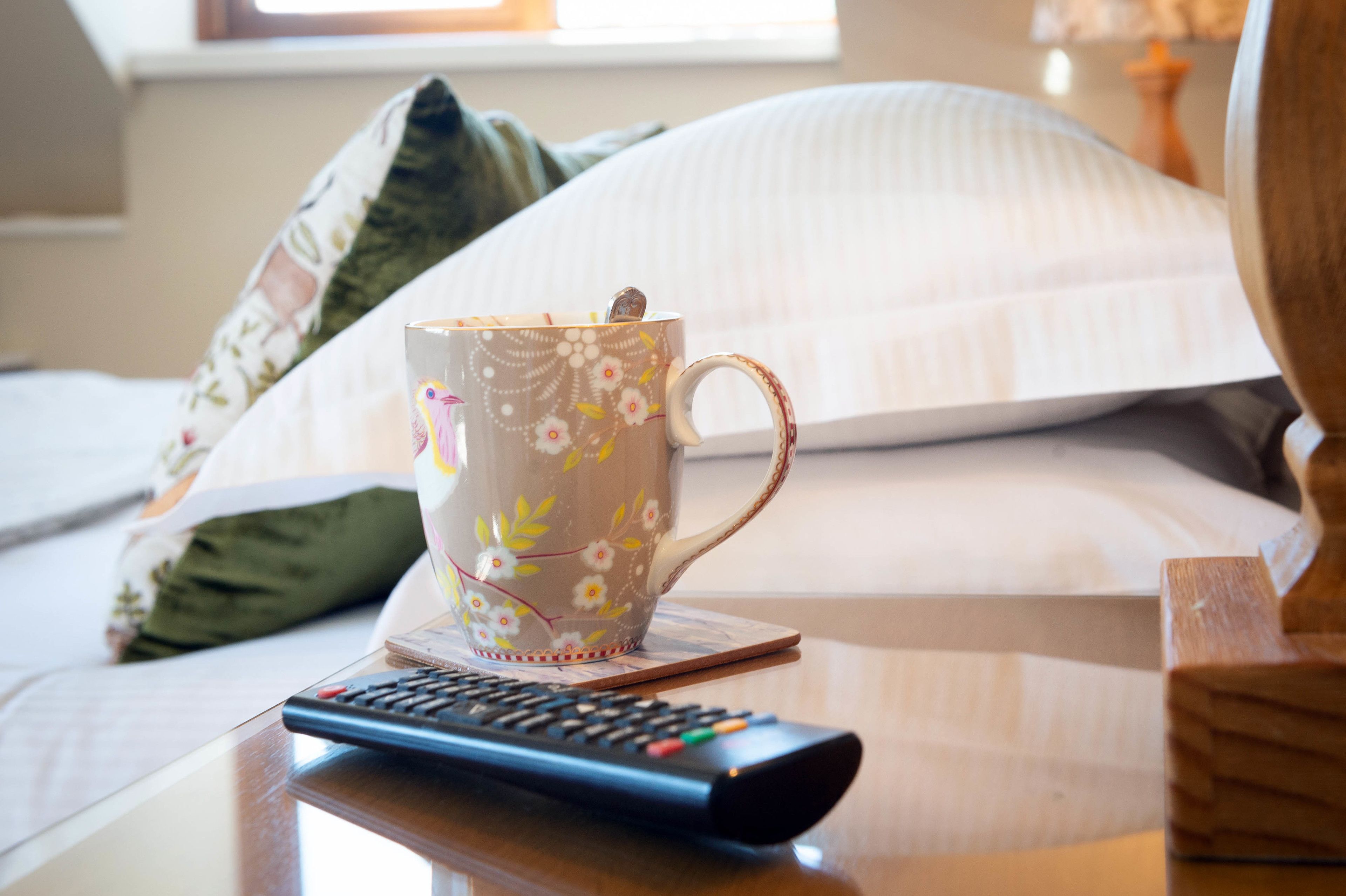 A bed with a floral-patterned mug and a remote control on a wooden table.