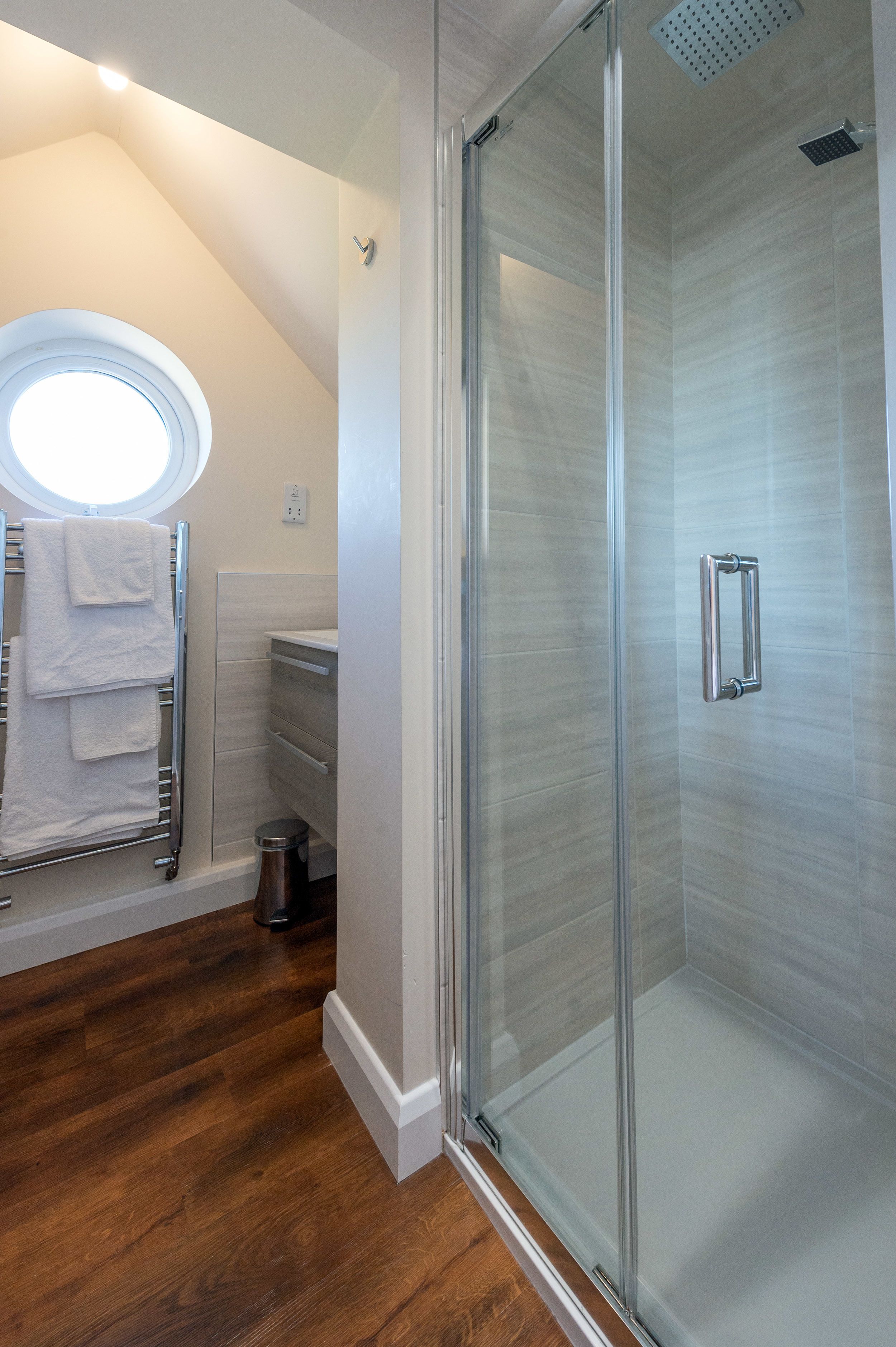 A modern bathroom with a glass-enclosed shower, a towel rack with white towels, and a wooden floor.