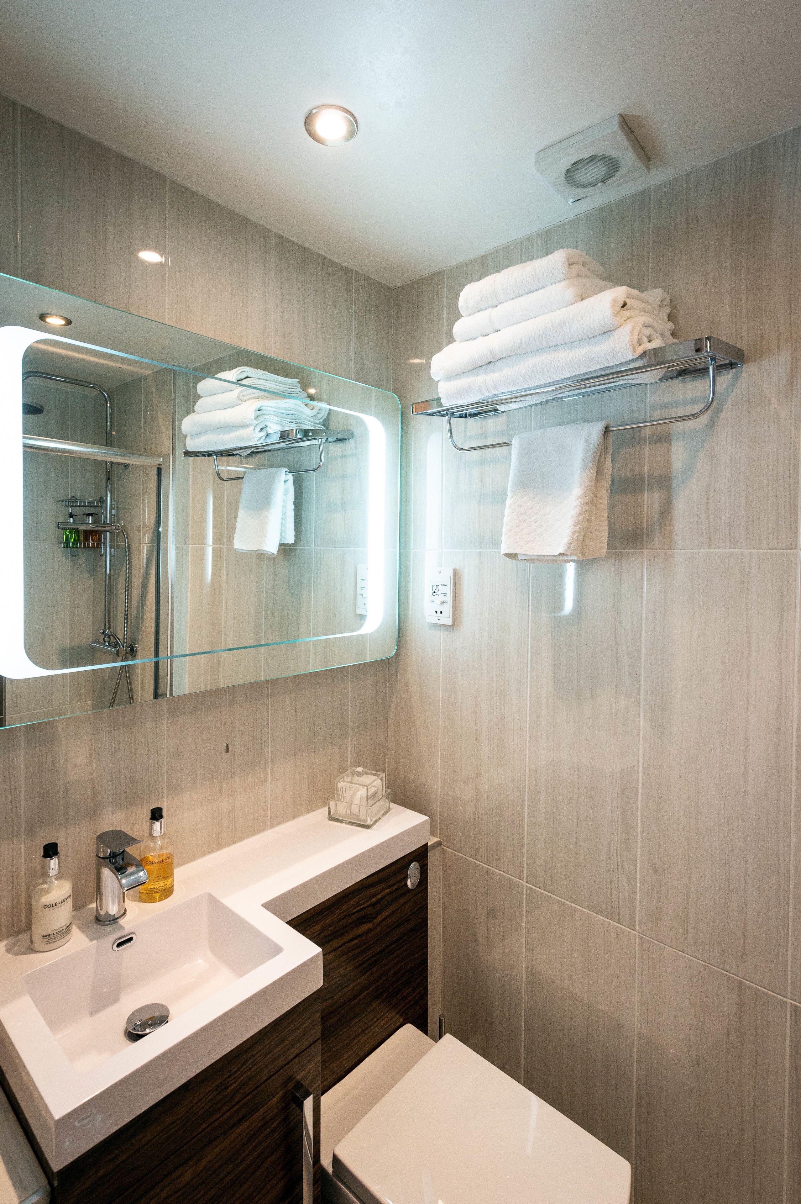 A modern bathroom with a large mirror, a glass shelf with towels, a white sink, and a shower area.