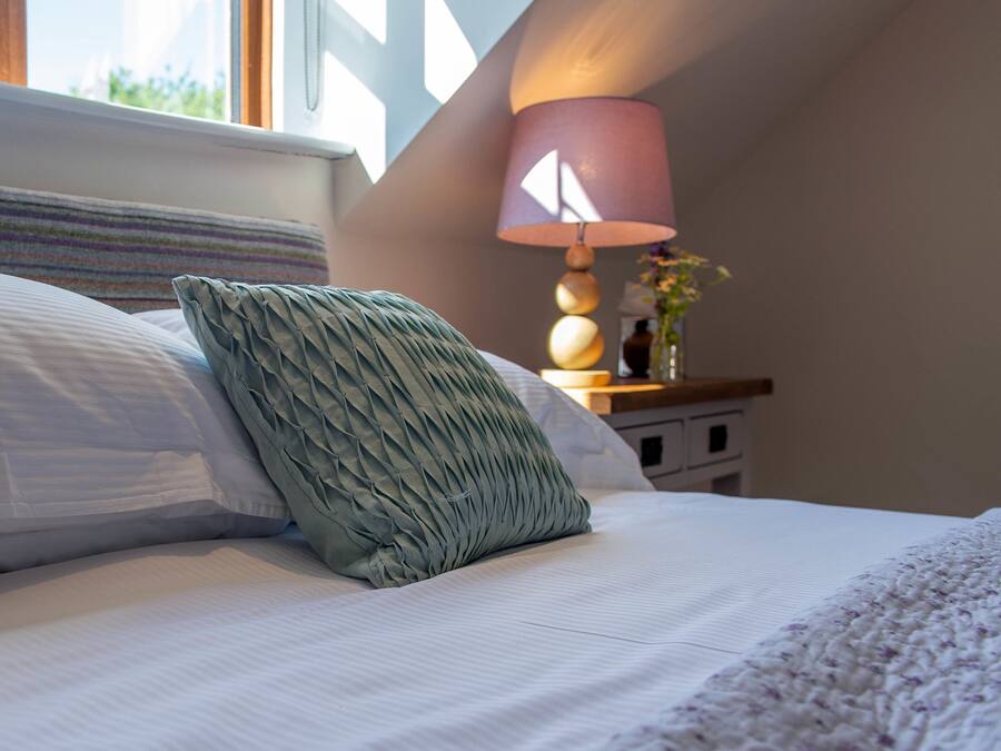 A neatly made bed with a striped headboard, a green textured pillow, and a white blanket. There is a bedside table with a lamp and a vase with flowers.