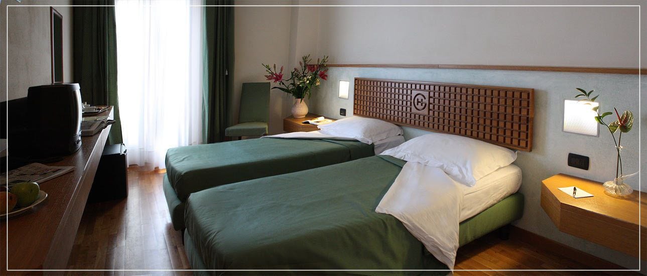 Une chambre d’hôtel avec deux lits, un bureau avec une chaise, une télévision, une lampe et un vase avec des fleurs.