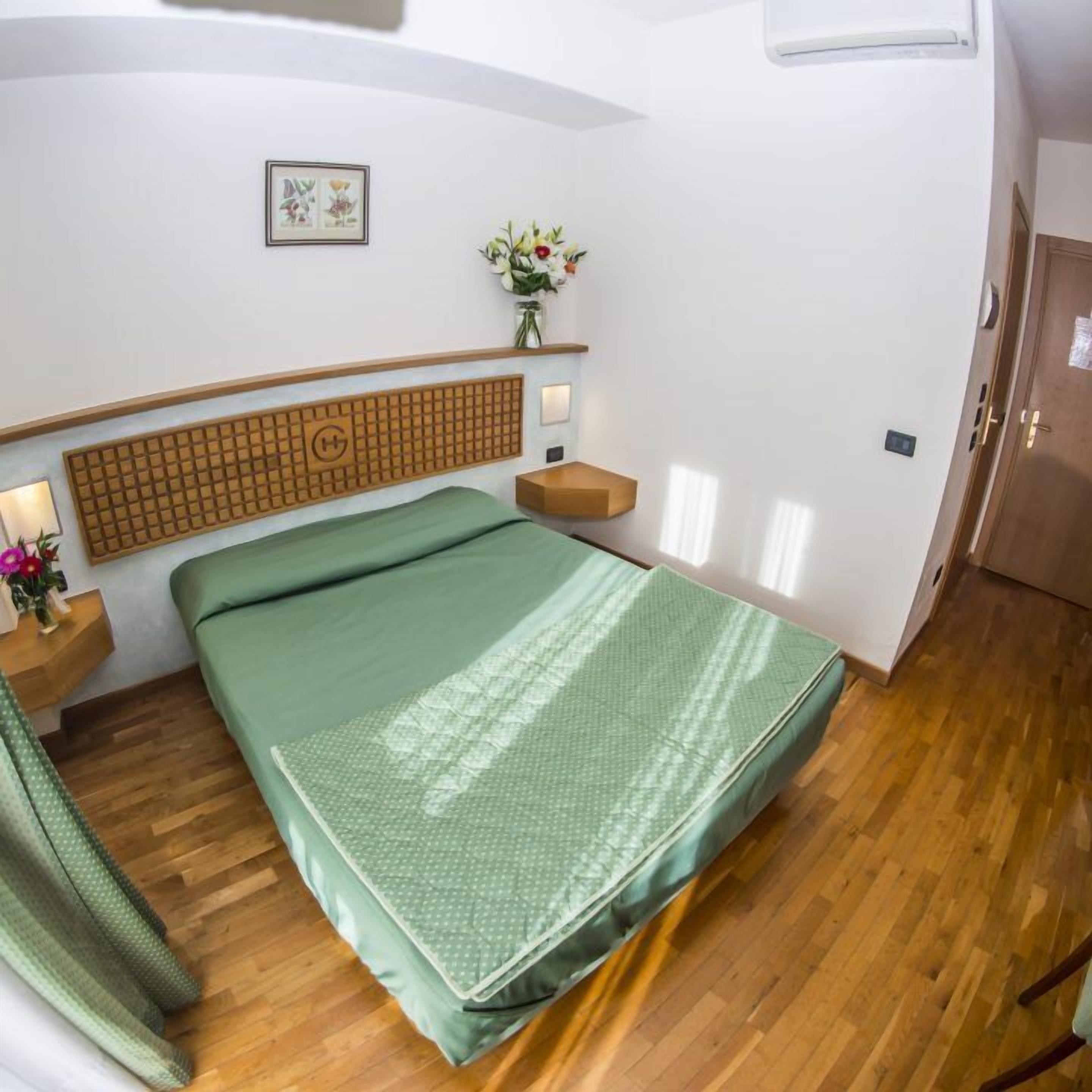 A compact hotel room with a single bed, a wooden headboard, a small bedside table with a lamp, a wall-mounted shelf with a vase of flowers, and a wooden wardrobe.