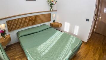 A compact hotel room with a single bed, a wooden headboard, a small bedside table with a lamp, a wall-mounted shelf with a vase of flowers, and a wooden wardrobe.