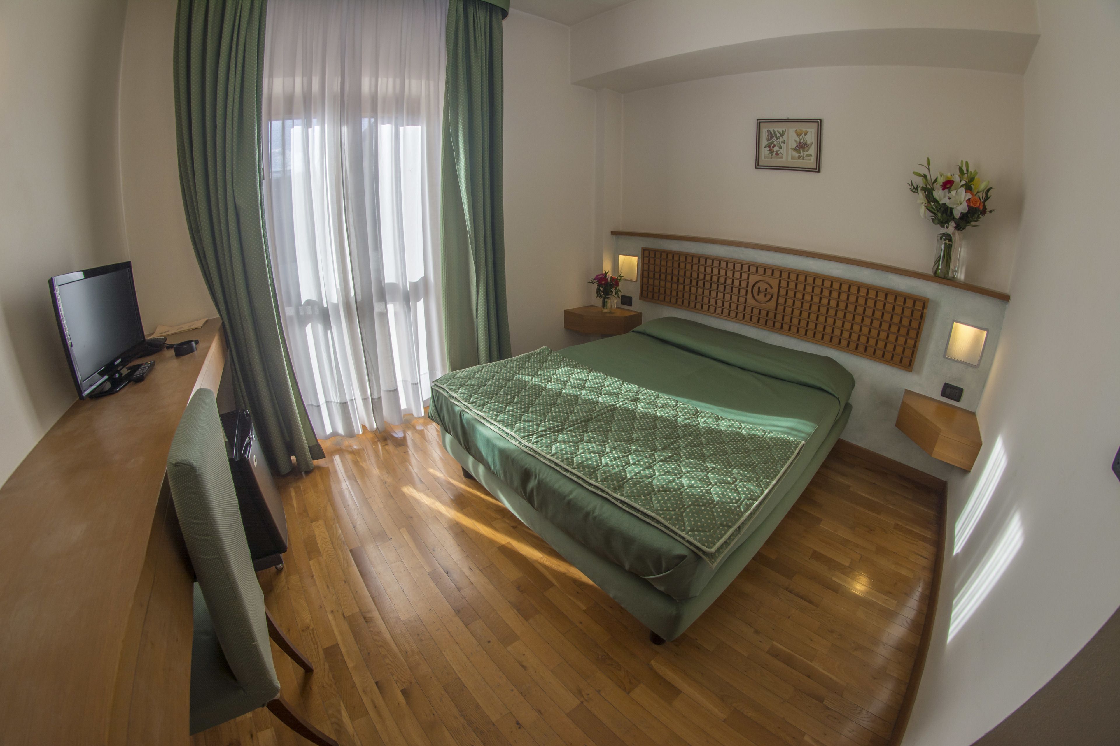 A hotel room with a bed, a desk with a computer, a TV, and a window with curtains.