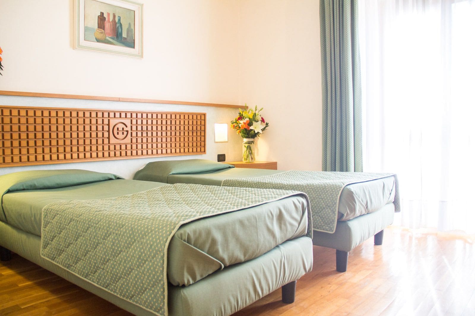 Habitación de hotel con dos camas, cabecero de madera, mesita de noche con un florero y un cuadro en la pared.