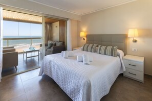 Ein Hotelzimmer mit einem großen Bett, zwei Nachttischlampen, Blick auf den Strand und einem Sitzbereich auf dem Balkon.