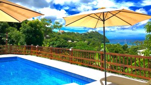 A swimming pool with a wooden deck and umbrellas, surrounded by lush greenery and a view of the ocean.