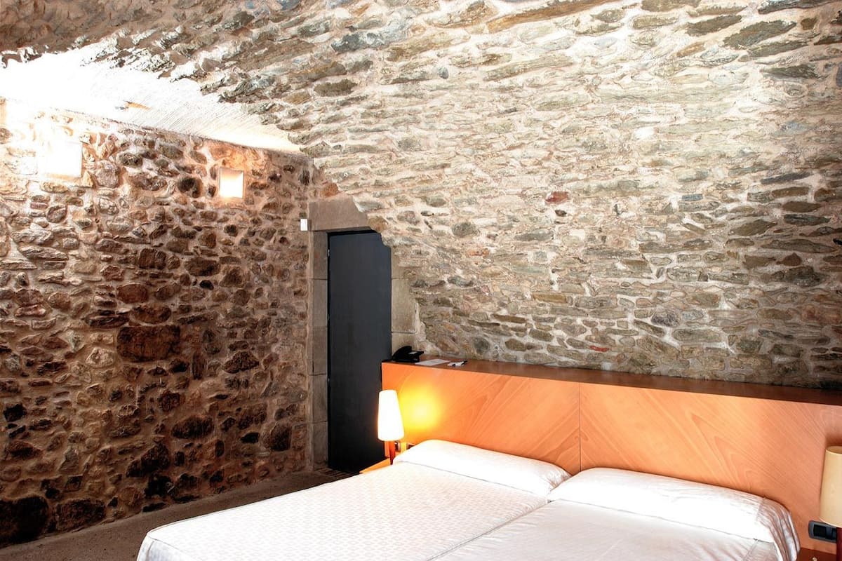 Una habitación con pared de piedra, cabecera de madera, cama con ropa blanca y mesita de noche con lámpara.