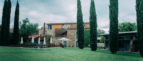 A courtyard with a stone building, tall cypress trees, and outdoor seating.