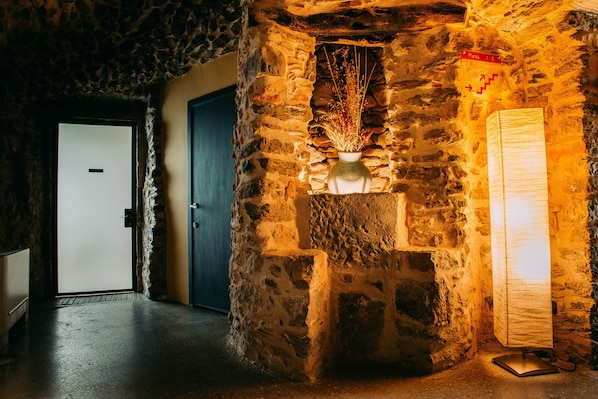 Un couloir aux murs de pierre, avec une porte, un vase contenant des plantes séchées et un lampadaire.