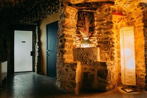 A stone-walled hallway with a door, a vase with dried plants, and a standing lamp.