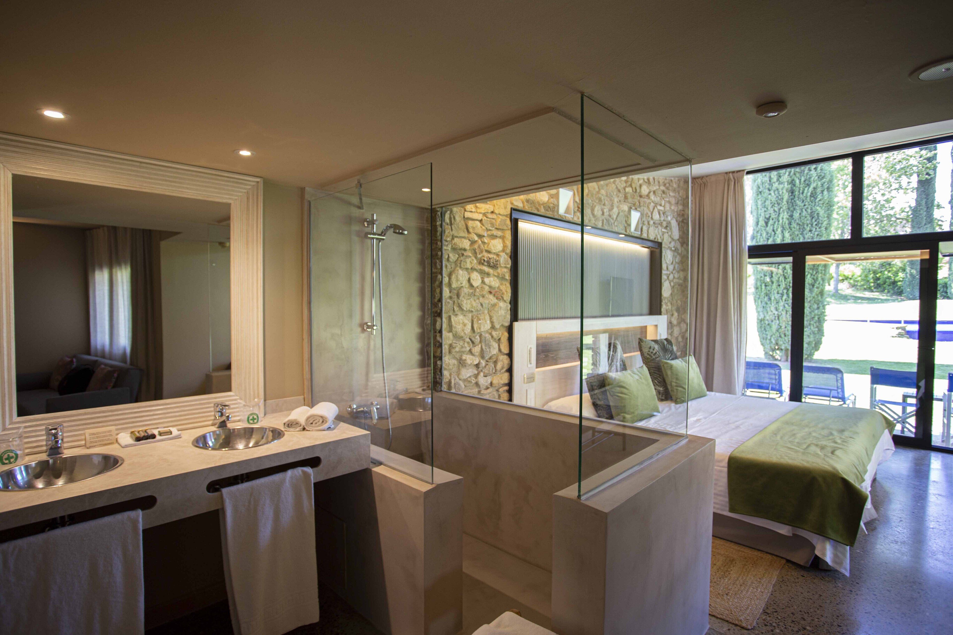 A modern bathroom with a large mirror, two sinks, a shower, and a bed with a green bedspread.