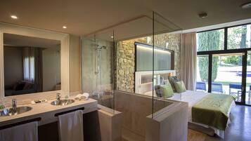 A modern bathroom with a large mirror, two sinks, a shower, and a bed with a green bedspread.
