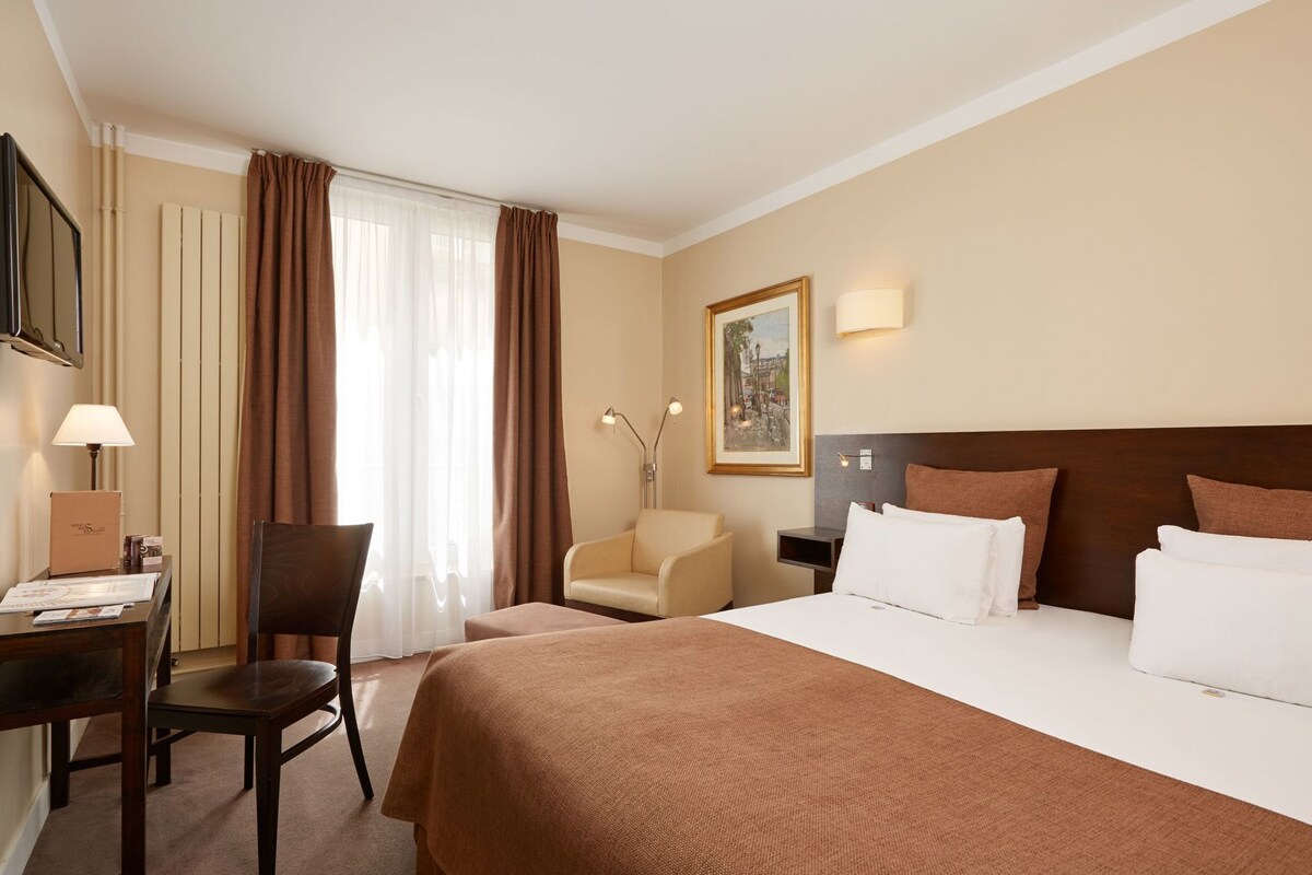 A hotel room with a bed, a desk, a chair, a lamp, and a painting on the wall.