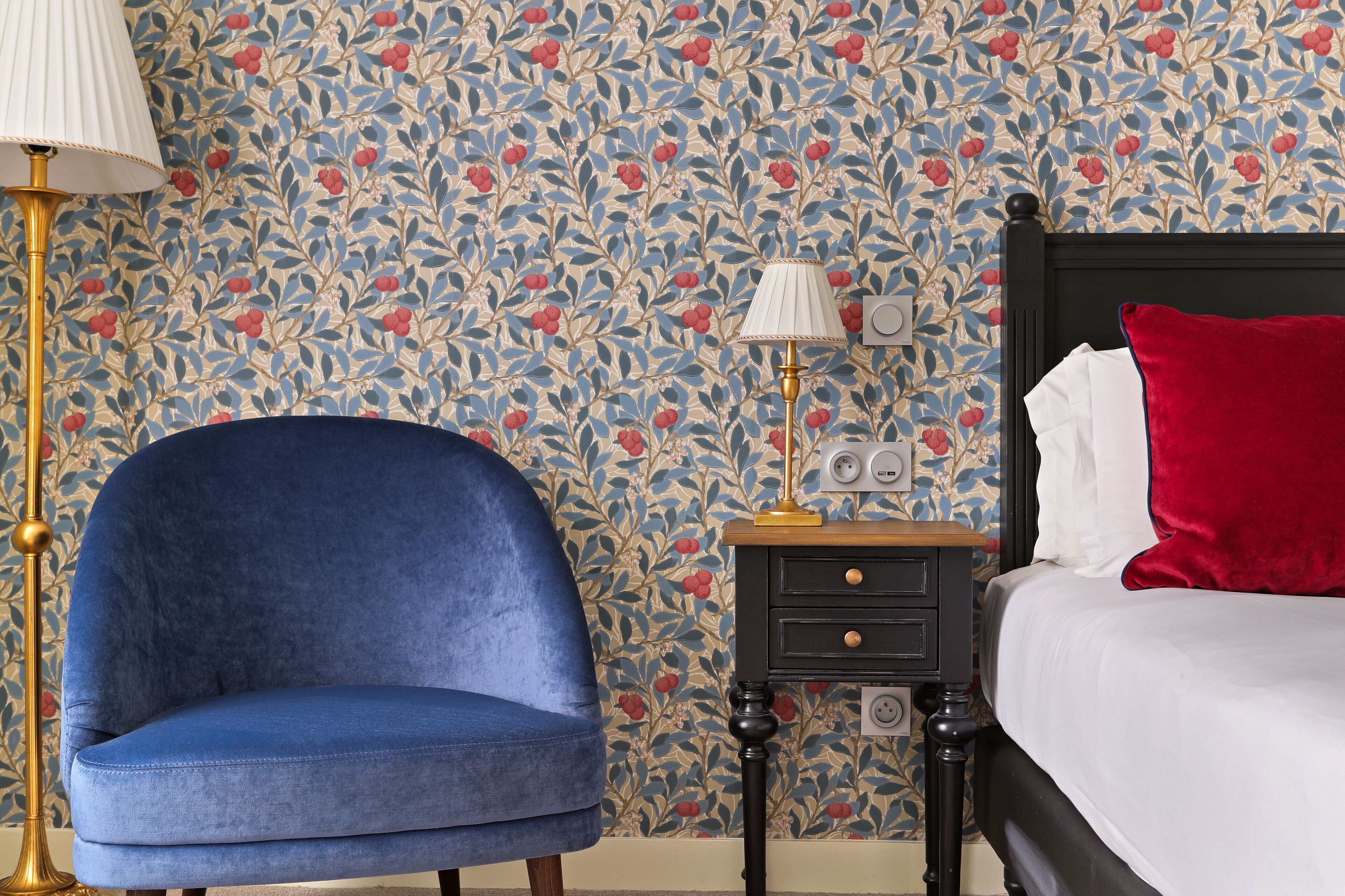 Une pièce avec un fauteuil bleu, une table de chevet avec une lampe et un lit avec un oreiller rouge.