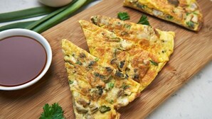 A wooden cutting board with several pieces of a savory pancake-like dish, garnished with green herbs, accompanied by a small bowl of dark sauce. - Hotel Indo continental (New Delhi)