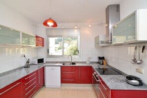 A modern kitchen with red cabinets, a built-in dishwasher, a stainless steel range hood, and a window providing natural light. - Lloret de Mar Villa: privacy, pool, nature, with breathtaking view (Lloret de Mar)