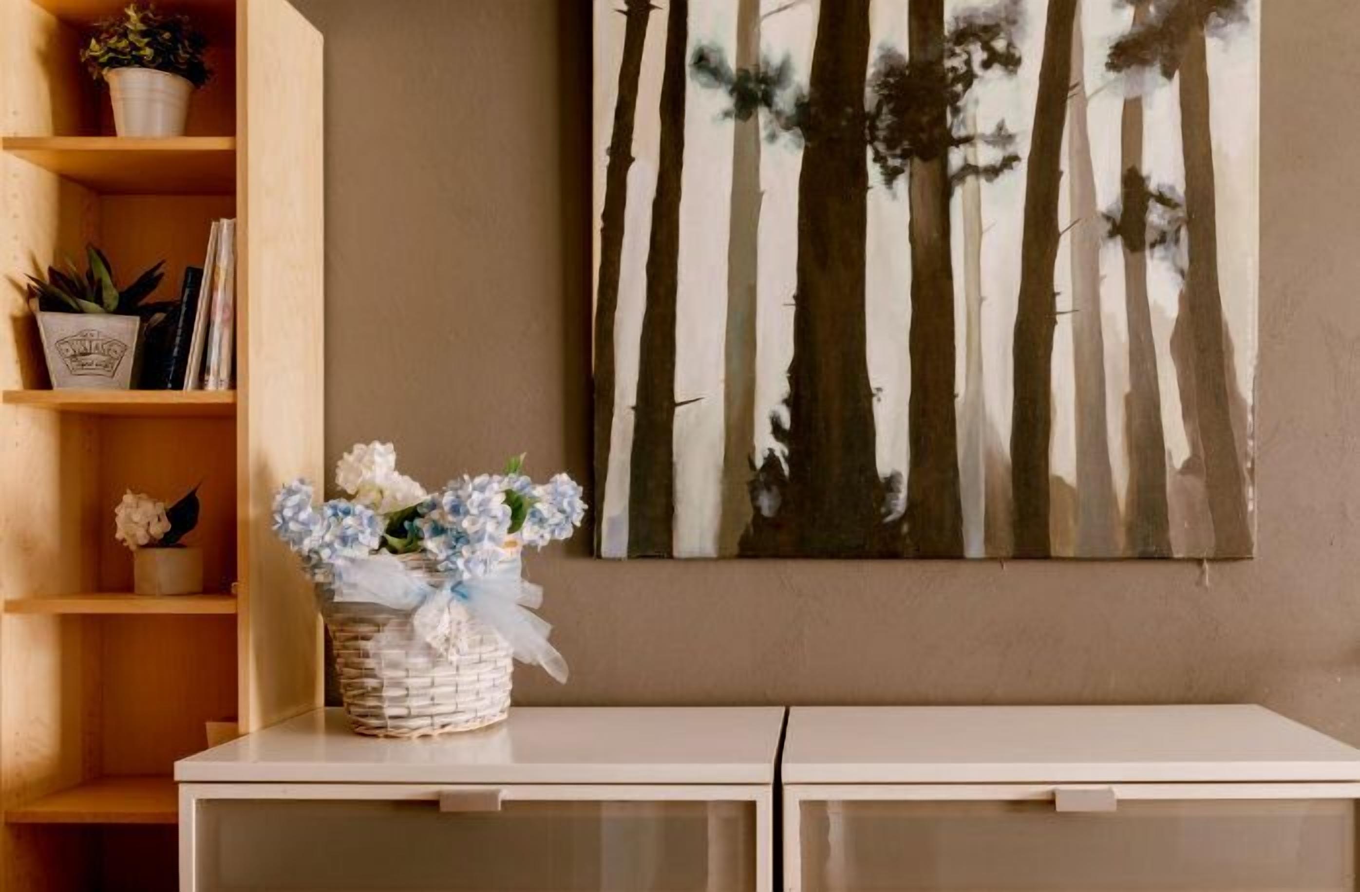 A room with a white console table, a wall-mounted painting of trees, and a shelf with plants and books.