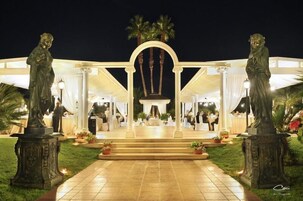 Espaço amplo para eventos ao ar livre, com palco central, flanqueado por duas estátuas, e uma galeria coberta que leva ao palco.
