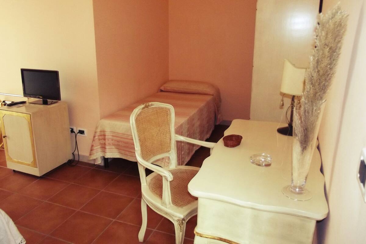 Une petite pièce, bien rangée, avec un lit, une chaise, un bureau avec un ordinateur et une lampe.