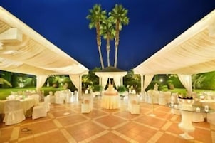 Um espaço para eventos ao ar livre com piso ladrilhado, mesas e cadeiras brancas, e uma estrutura decorativa central.