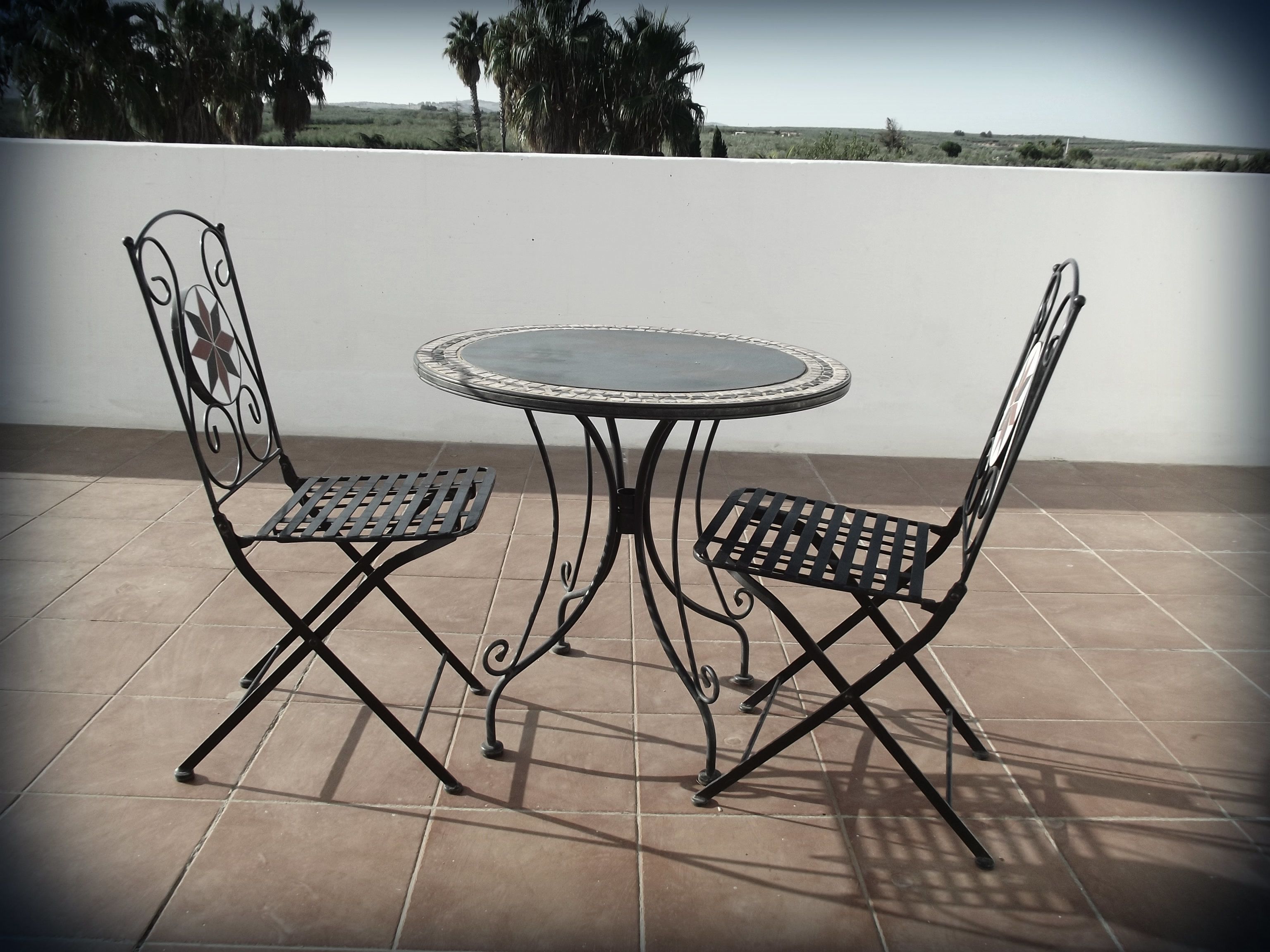 A patio with a round table and two chairs, a white wall, and a view of the landscape.