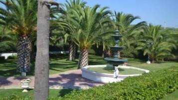 A well-maintained garden with a central fountain, surrounded by palm trees and neatly trimmed hedges.