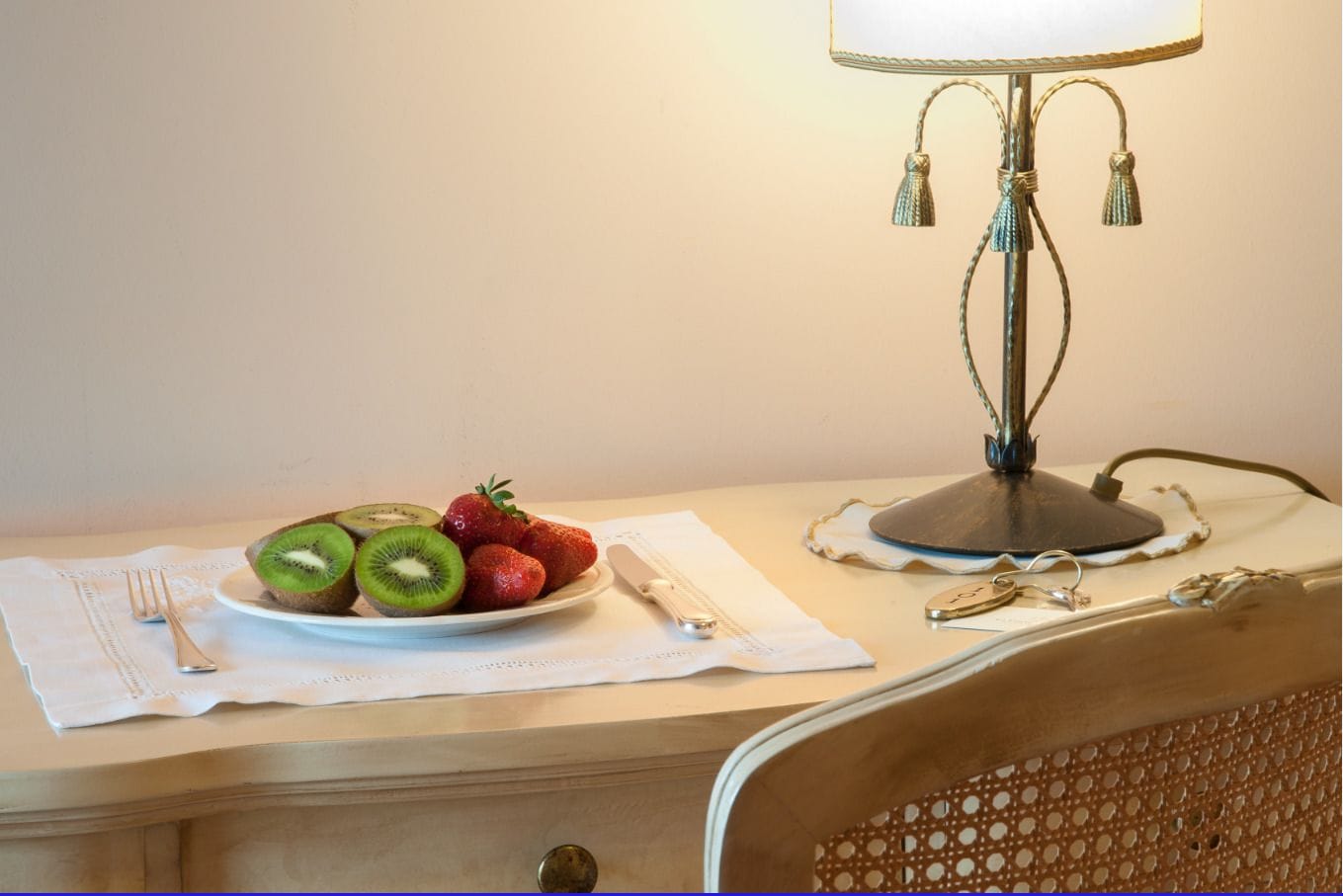 A dining table with a plate of sliced kiwi and strawberries, a fork, a knife, and a lamp.
