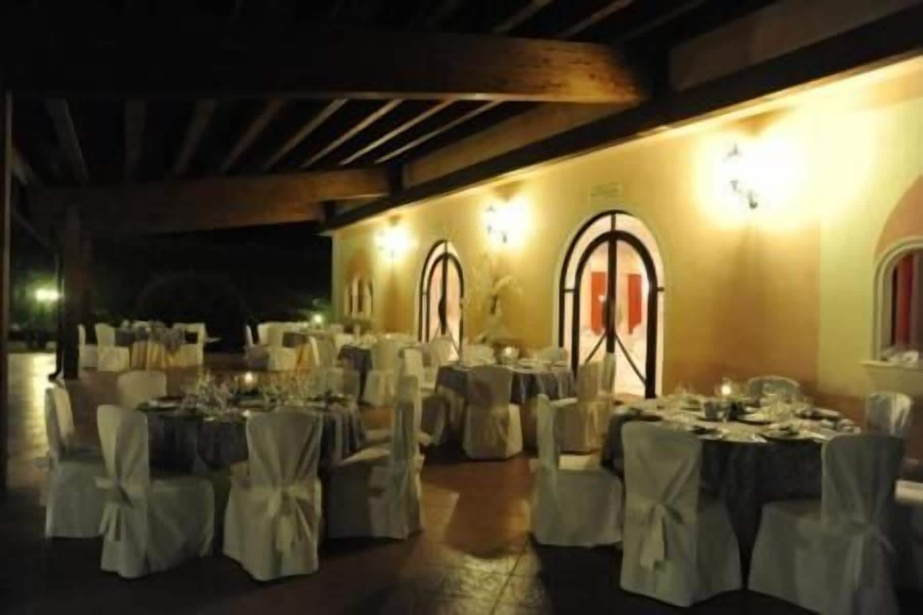 Un salón de banquetes al aire libre con mesas redondas cubiertas con manteles blancos y sillas, iluminado por luces fijadas en la pared.