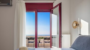 Ett hotellrum med en säng, en hatt på sängen, utsikt över havet genom en öppen dörr och en balkong med stolar och ett bord.