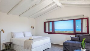 Ett sovrum med en säng, ett sängbord, en lampa, en soffa, ett fönster med utsikt över havet och en krukväxt.