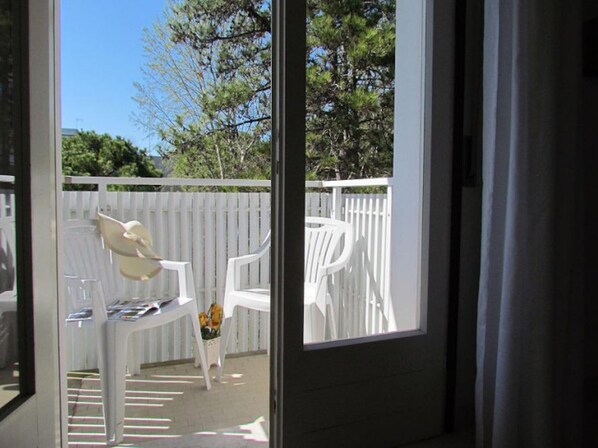 A view through a glass door to an outdoor patio area with white plastic chairs and a table, a white fence, and trees in the background.
