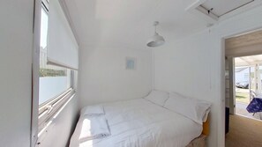 Una habitación pequeña con cama individual, paredes blancas, ventana, una lámpara en el techo y puerta que da al exterior.