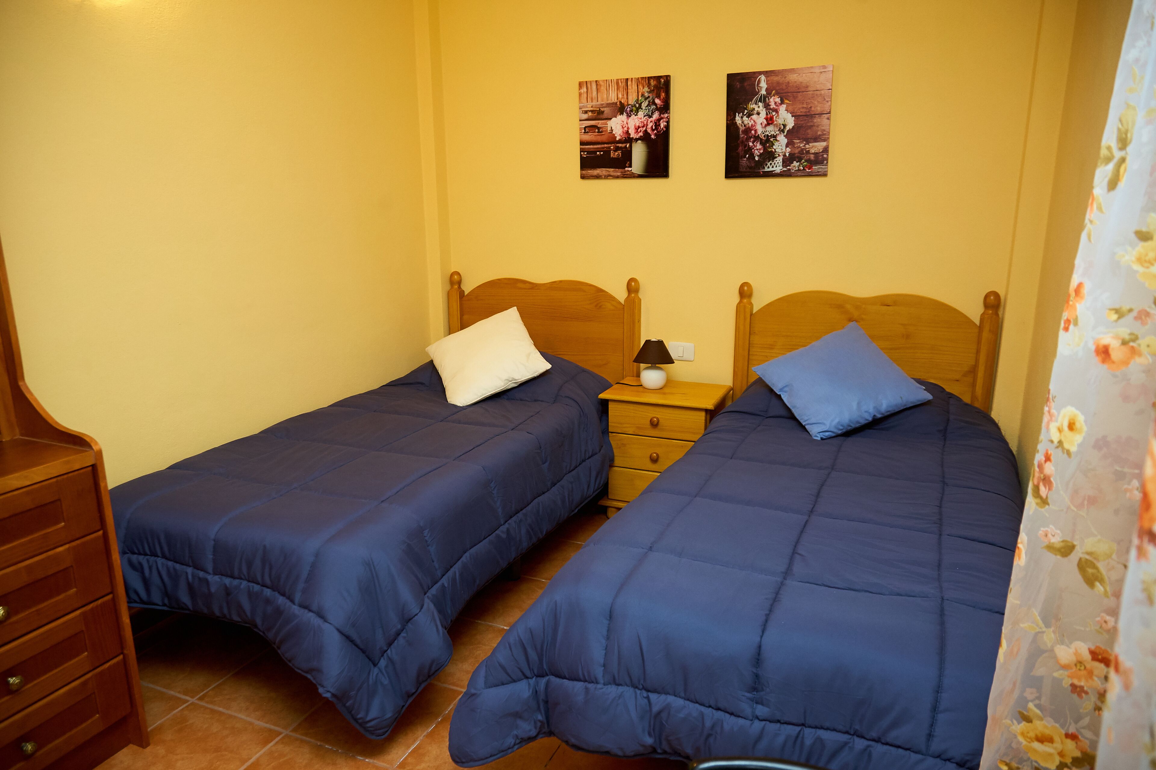 Een kamer met twee eenpersoonsbedden, houten hoofdeinden, een nachtkastje met lamp en een houten dressoir.