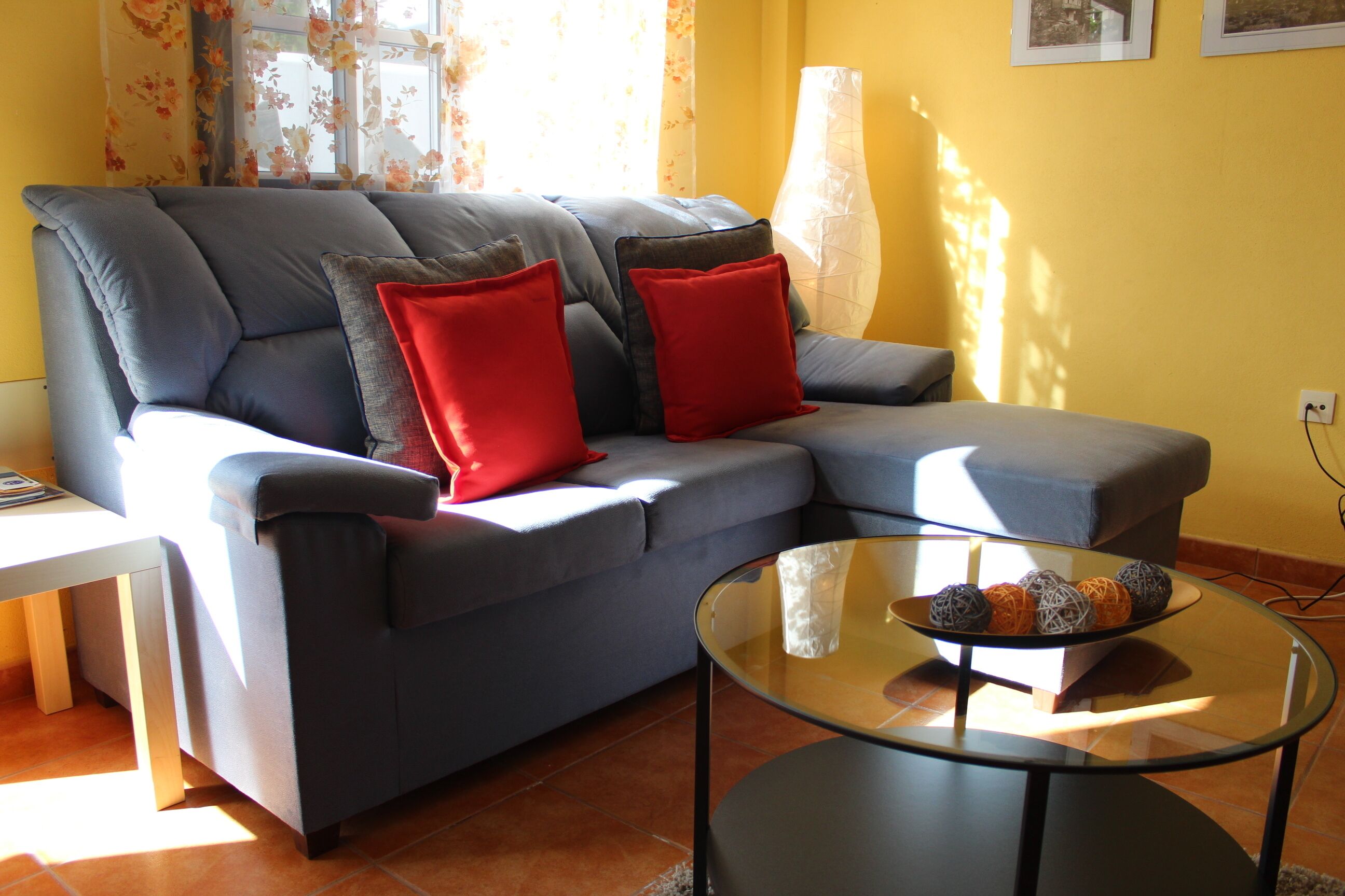 A living room with a grey sofa, red cushions, a glass coffee table with decorative balls, and a white side table.